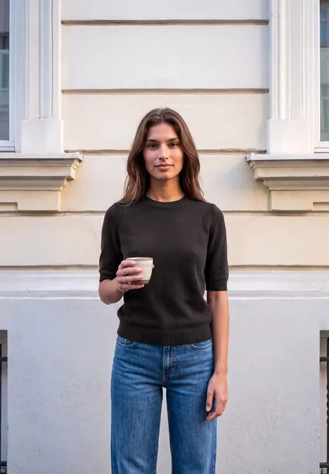 Sweter z krótkim rękawem i okrągłym dekoltem charred brown mel. Młoda kobieta stoi przy elewacji, trzyma kubek kawy, ma na sobie czarny T-shirt i dżinsy.