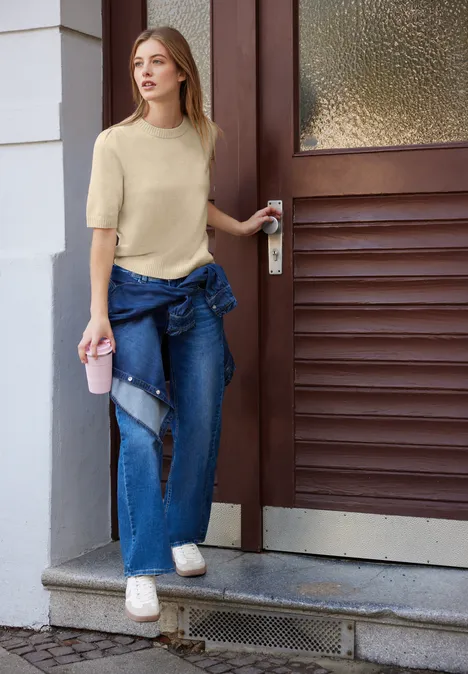 Frau mit beigem T-Shirt, zerrissenen Jeans und Denim-Jacke um die Hüfte hält einen Becher und öffnet eine Haustür.