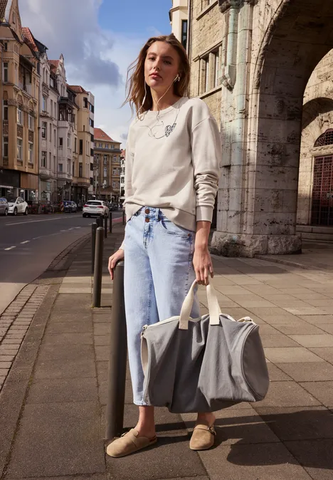 Person mit verschwommenem Gesicht in Jeans und Pulli steht vor historischen Häusern und hält große Tasche.