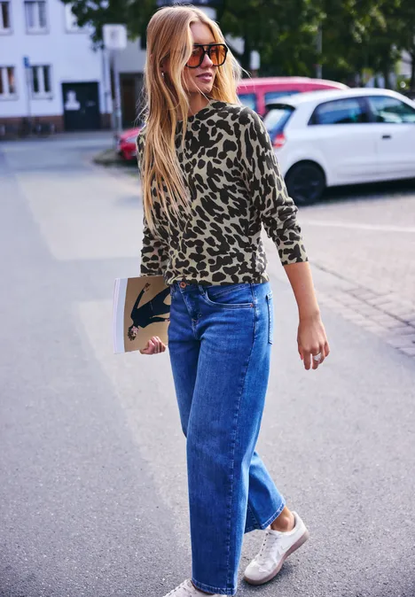 Eine Frau mit blondem Haar in Animal-Print-Shirt, Jeans und Sneakers läuft auf der Straße und hält eine Clutch.