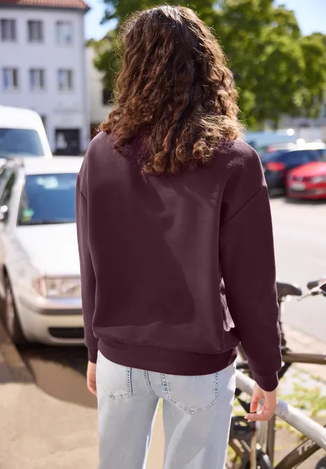 Person von hinten in bordeauxrotem Sweatshirt und heller Jeans, die auf einem sonnigen Bürgersteig entlangläuft.
