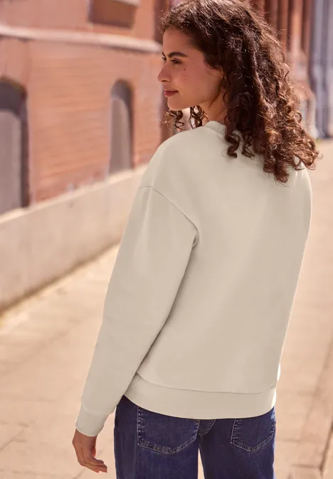 Frau mit lockigem Haar in beigefarbenem Pullover und Jeans steht seitlich auf einem sonnigen Gehweg.