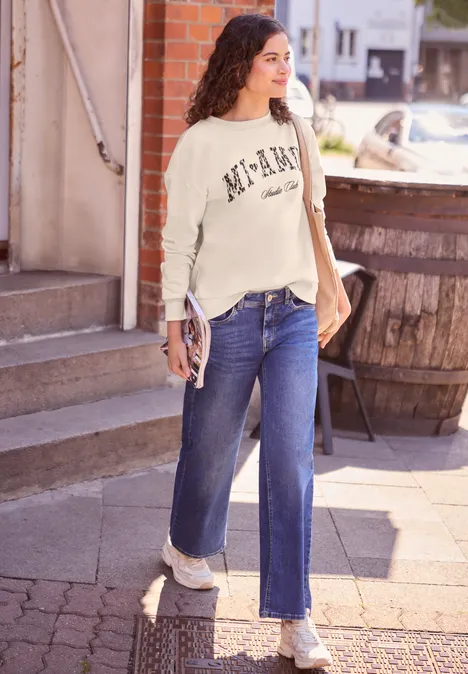Frau trägt Jeans, Pulli und Turnschuhe, geht auf gepflastertem Weg, hält Tasche und Hefter.
