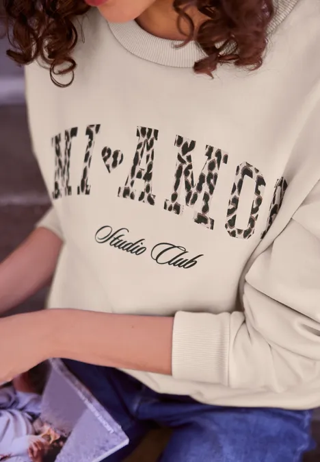 Person trägt hellbeiges Sweatshirt mit Schriftzug "NI’AMOR Studio Club" und hält ein Buch auf dem Schoß.