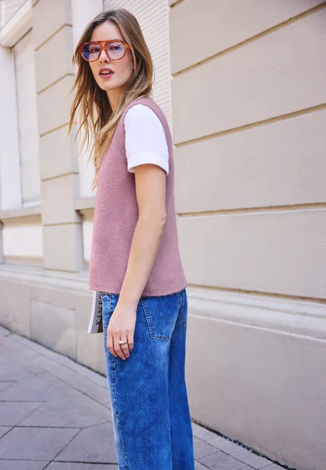 Frau mit rosa Pullunder, weißem T-Shirt, blauen Jeans und Brille steht auf einem Gehweg vor einem Gebäude.