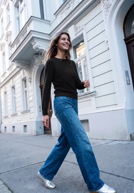 Junge Frau in schwarzem Pullover und blauen Jeans spaziert selbstbewusst eine Straßenfassade entlang.