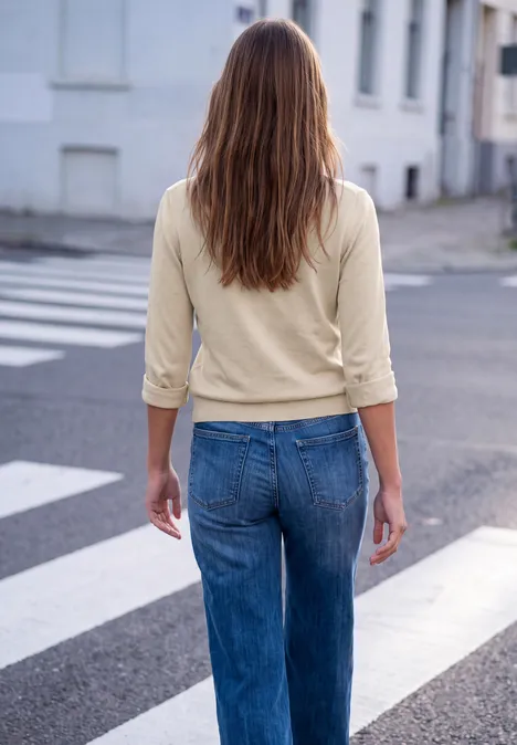 Frau mit langen braunen Haaren in beiger Bluse und blauen Jeans überquert einen Zebrastreifen.