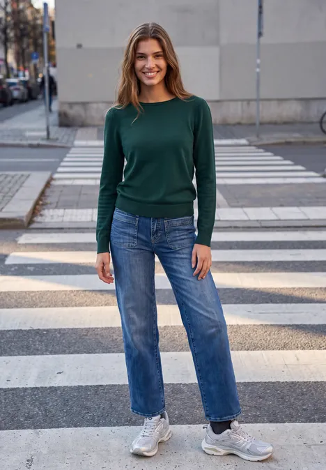Frau mit grünem Pullover, blauen Jeans und Sneakers lächelt auf einem Zebrastreifen in der Stadt.