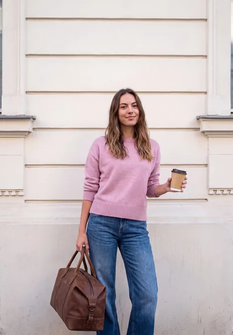 Junge Frau in Jeans und rosafarbenem Pullover mit Tasche und Kaffee vor heller Hauswand.