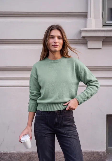 Pullover mit Rundhals und Knopfdetails grayed mint mel. Frau mit grünem Pullover und dunklen Jeans steht vor einem Gebäude und hält einen Kaffeebecher.