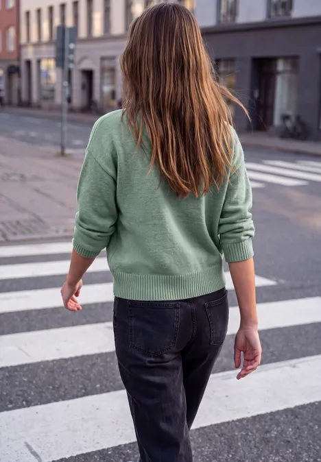 Pullover mit Rundhals und Knopfdetails grayed mint mel. Eine Frau mit grünem Pullover und Jeans überquert von hinten einen Zebrastreifen in einer Stadt.