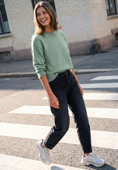 Pullover mit Rundhals und Knopfdetails grayed mint mel. Frau in grünem Pullover läuft fröhlich über Zebrastreifen auf Asphaltstraße, trägt Jeans und weiße Sneaker.
