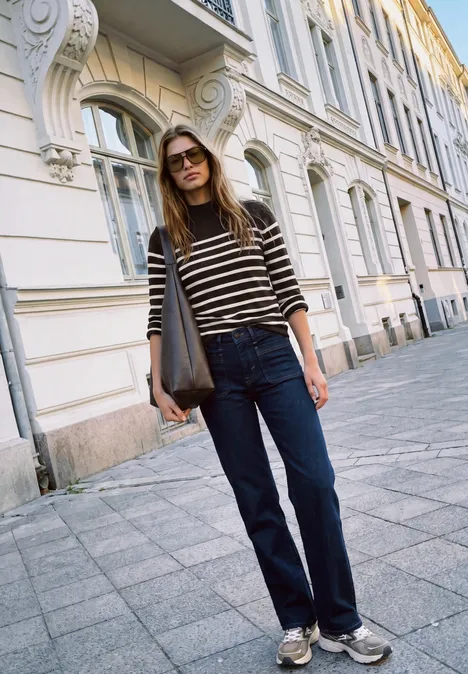Sweter z golfem i w paski charred brown Młoda kobieta w pasiastym swetrze, ciemnych jeansach i trampkach z dużą torbą, swobodnie stojąca przed białą kamienicą.