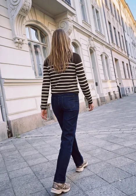 Sweter z golfem i w paski charred brown Osoba w pasiastym swetrze i dżinsach idzie chodnikiem obok eleganckiej kamienicy.