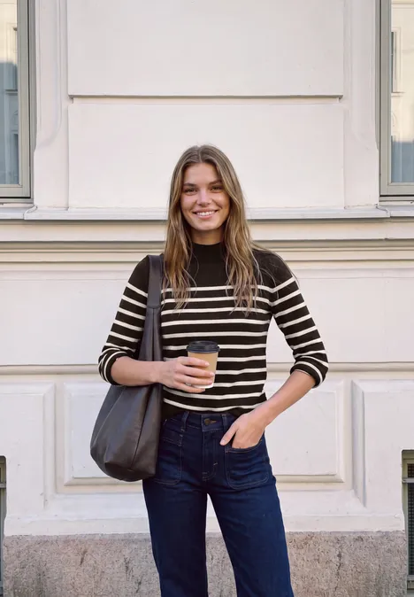 Sweter z golfem i w paski charred brown Młoda kobieta uśmiecha się przed fasadą, trzyma kawę na wynos i nosi pasiasty sweter oraz dżinsy.