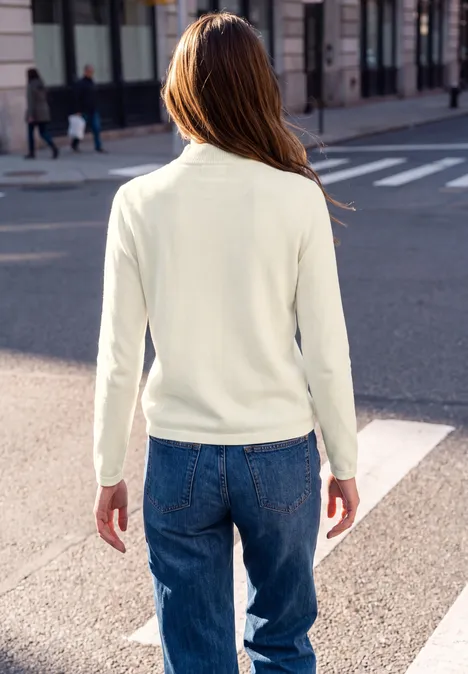 Sweter z długim rękawem i golfem off white Osoba widziana od tyłu w białym swetrze i niebieskich dżinsach przechodzi przez ulicę.