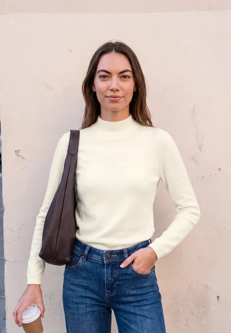 Sweter z długim rękawem i golfem off white Kobieta w białym golfie i dżinsach z brązową torbą na ramieniu trzyma papierowy kubek przed beżową ścianą.