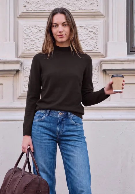 Sweter z długim rękawem i golfem charred brown Młoda kobieta w dżinsach i swetrze trzyma kawę i torbę przed zabytkową elewacją.