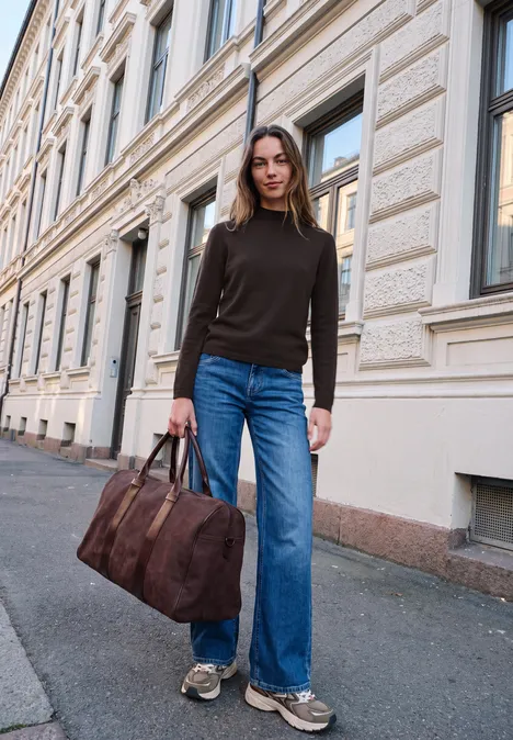 Sweter z długim rękawem i golfem charred brown Kobieta idzie obok eleganckiej fasady, w czarnym golfie, szerokich dżinsach i z brązową torbą podróżną.