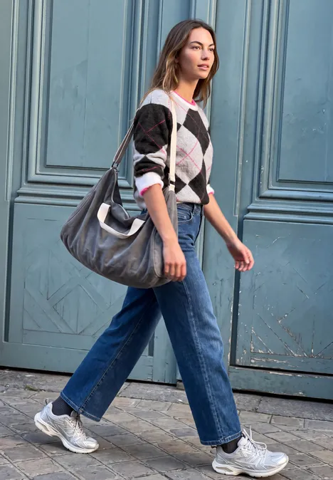Sweter z okrągłym dekoltem i wzorem argyle off white Kobieta w dżinsach, swetrze i trampkach z dużą torbą idzie obok niebieskich drzwi.