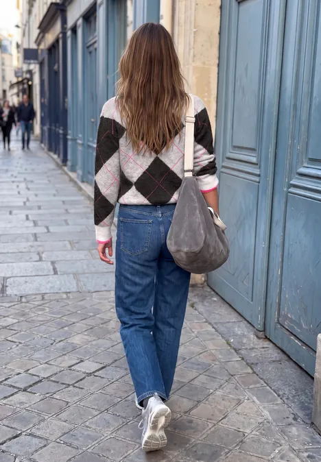 Sweter z okrągłym dekoltem i wzorem argyle off white Kobieta w dżinsach, swetrze w romby i białych trampkach spaceruje po brukowanej ulicy.