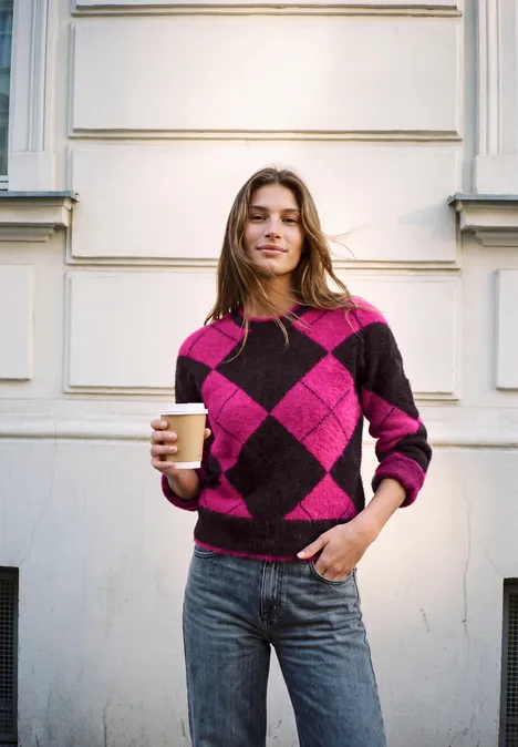 Sweter z okrągłym dekoltem i wzorem argyle charred brown Młoda kobieta w różowo-fioletowym swetrze w romby, trzyma kubek kawy przed białą ścianą.