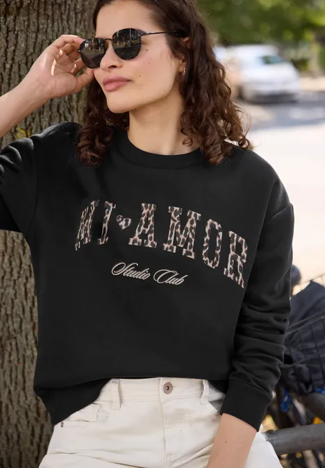 Junge Frau mit Sonnenbrille und schwarzem Pullover mit Leoparden-Aufdruck steht lässig an einem Baum.
