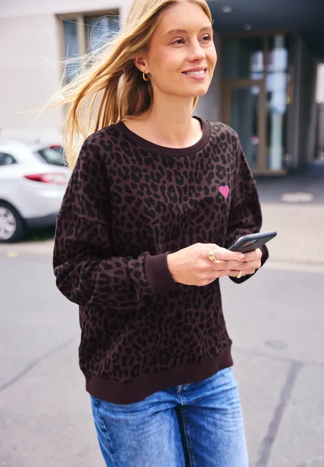 Kobieta w dżinsach i leopardim swetrze idzie ulicą, uśmiecha się i trzyma smartfon.