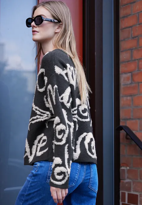 Junge Frau mit Sonnenbrille trägt schwarzen Pullover mit weißem Schriftzug und blaue Jeans.