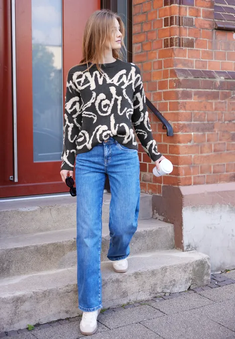 Frau verlässt ein Backsteinhaus mit Kaffebecher und Schlüssel, trägt schwarzen Pullover und blaue Jeans.