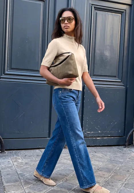 Femme portant des lunettes, un pull beige, un jean bleu et un sac, marchant devant une porte sombre