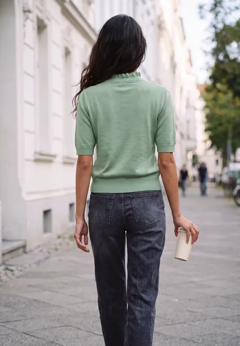 Femme en pull vert et jean, tasse à café à la main, marche en ville.
