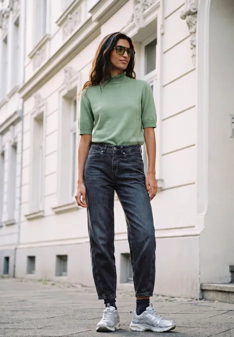Femme en pull vert et jean debout sur le trottoir devant un immeuble.