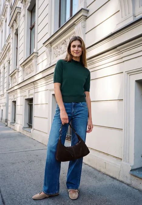 Pull à manches courtes et col roulé mossed green Eine lächelnde Frau in grünem Oberteil, blauen Jeans und beigen Flats steht mit Handtasche vor Hausfassade.