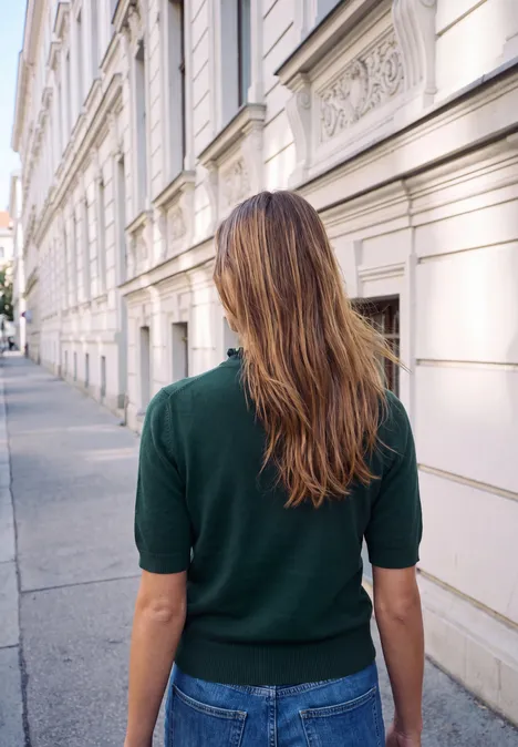 Pull à manches courtes et col roulé mossed green Frau mit braunem Haar und grünem Pullover geht an einer eleganten Häuserzeile vorbei.