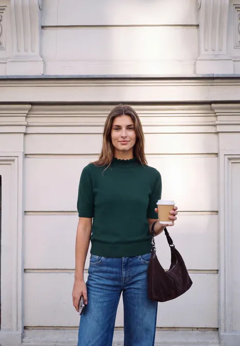 Pull à manches courtes et col roulé mossed green Junge Frau in grünem Pullover, Jeans, hält einen Kaffee und ein Handy vor einer hellen Hauswand.