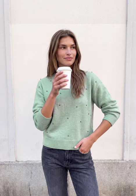 Trui met ronde hals en parelversieringen grayed mint mel. Vrouw in groene trui en donkere spijkerbroek, houdt koffiebeker vast, staat voor een lichte muur.