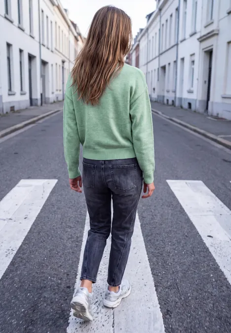 Trui met ronde hals en parelversieringen grayed mint mel. Een vrouw met een groen truitje, donkere spijkerbroek en witte sneakers steekt een rustige woonstraat over.