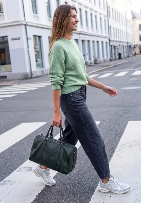Trui met ronde hals en parelversieringen grayed mint mel. Vrouw loopt over zebrapad in de stad, draagt groene trui, jeans, witte sneakers en een groene tas.