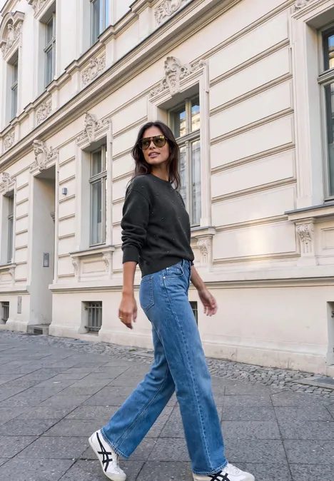 Frau in schwarzem Pullover, Jeans und weißen Sneakern spaziert vor historischem Gebäude.