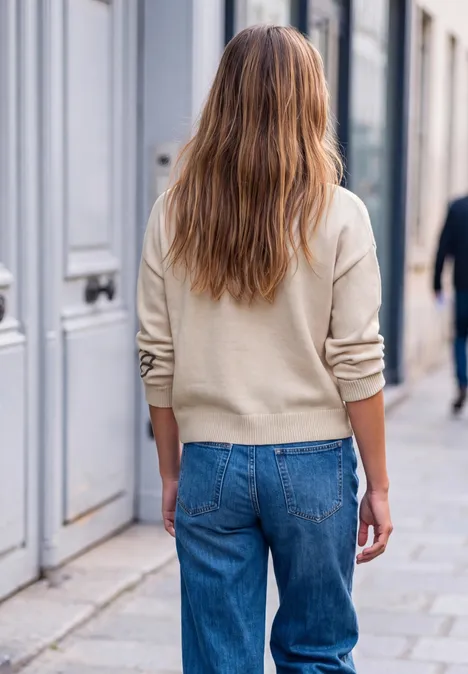 Frau mit beigem Pullover und blauer Jeans, von hinten gesehen, spaziert an einem städtischen Bürgersteig.