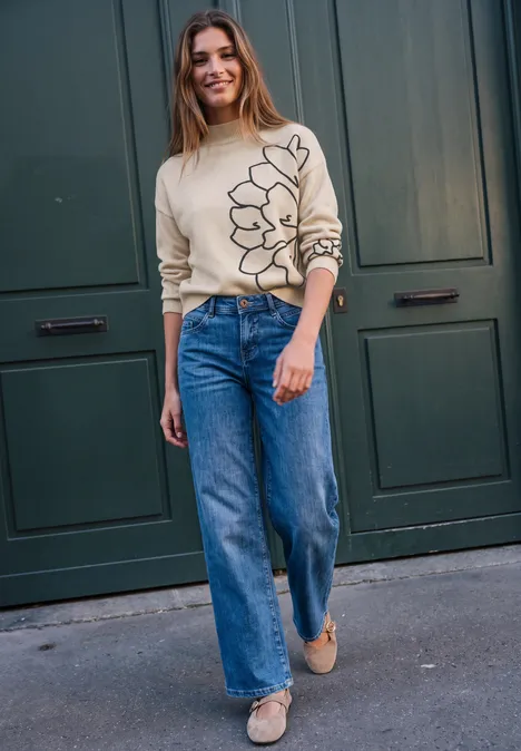 Junge Frau in beigem Pullover mit Blumenmotiv und weiten Jeans steht lächelnd vor einer grünen Tür.