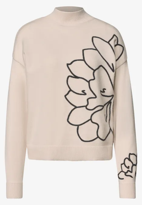 Beiger Rollkragenpullover mit stilisiertem schwarzen Blumenmotiv auf Vorderseite und Ärmel.