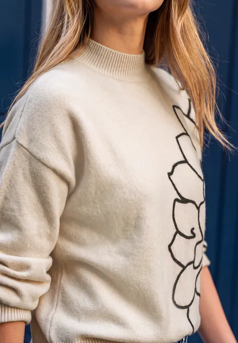 Frau im beigen Strickpullover mit schwarzem Blumenmuster vor dunkelblauem Hintergrund.