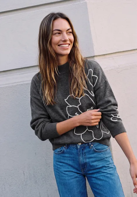 Pullover mit Turtle Neck und Stickerei inked grey mel. Lächelnde Frau mit grauem Blumensweater und blauen Jeans lehnt entspannt vor einer hellen Wand.