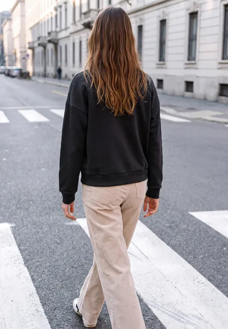 Persoon met lang haar, donkere trui en beige broek loopt over een zebrapad in een rustige stadsstraat.