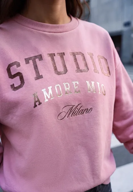 Frau trägt rosa Sweatshirt mit Aufdruck "STUDIO AMORE MIO Milano" vor urbanem Hintergrund