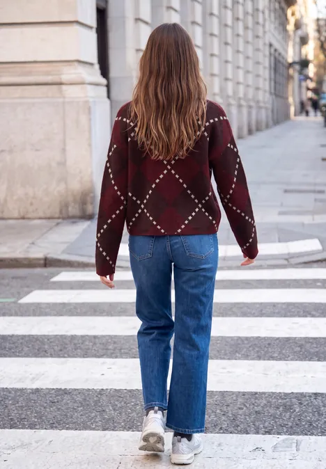 Eine Frau mit rotem Rautenmuster-Pullover, blauen Jeans und weißen Sneakern geht über einen Zebrastreifen in der Stadt.