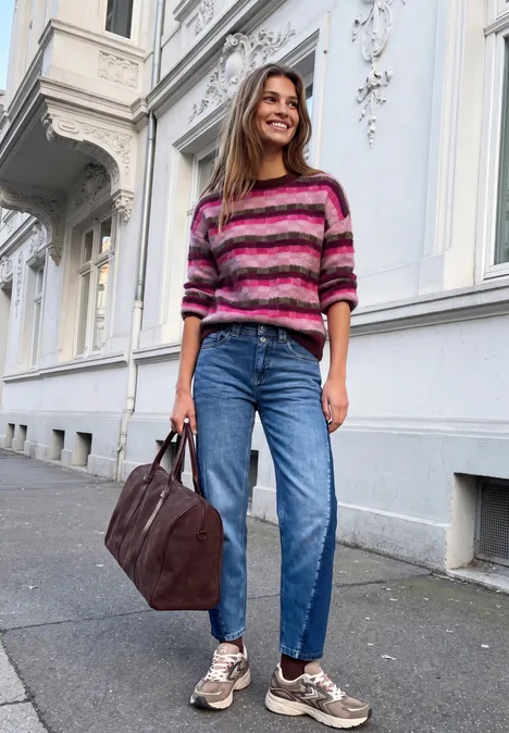 Eine junge Frau in gestreiftem Pullover, Jeans und Sneakers trägt eine Tasche und lächelt auf der Straße.
