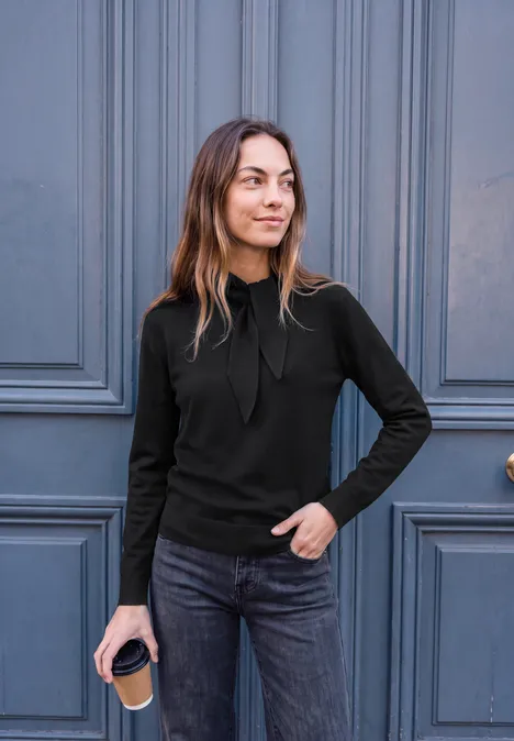 Langarm Pullover mit Schalkragen Black Junge Frau in lässigem Outfit hält einen Kaffeebecher vor einer blauen Tür.